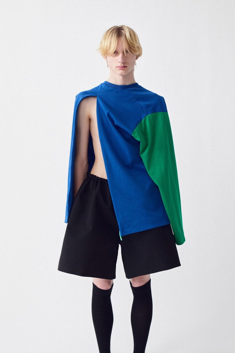 Young male model wearing an asymmetrical blue and green top with black shorts against a plain white background