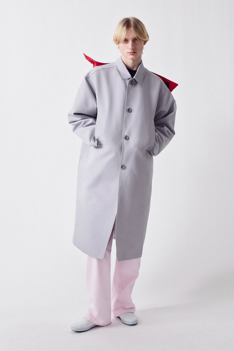 Full shot of a person in a light grey coat and pink pants standing in a bright white setting, fashion forward clothing.