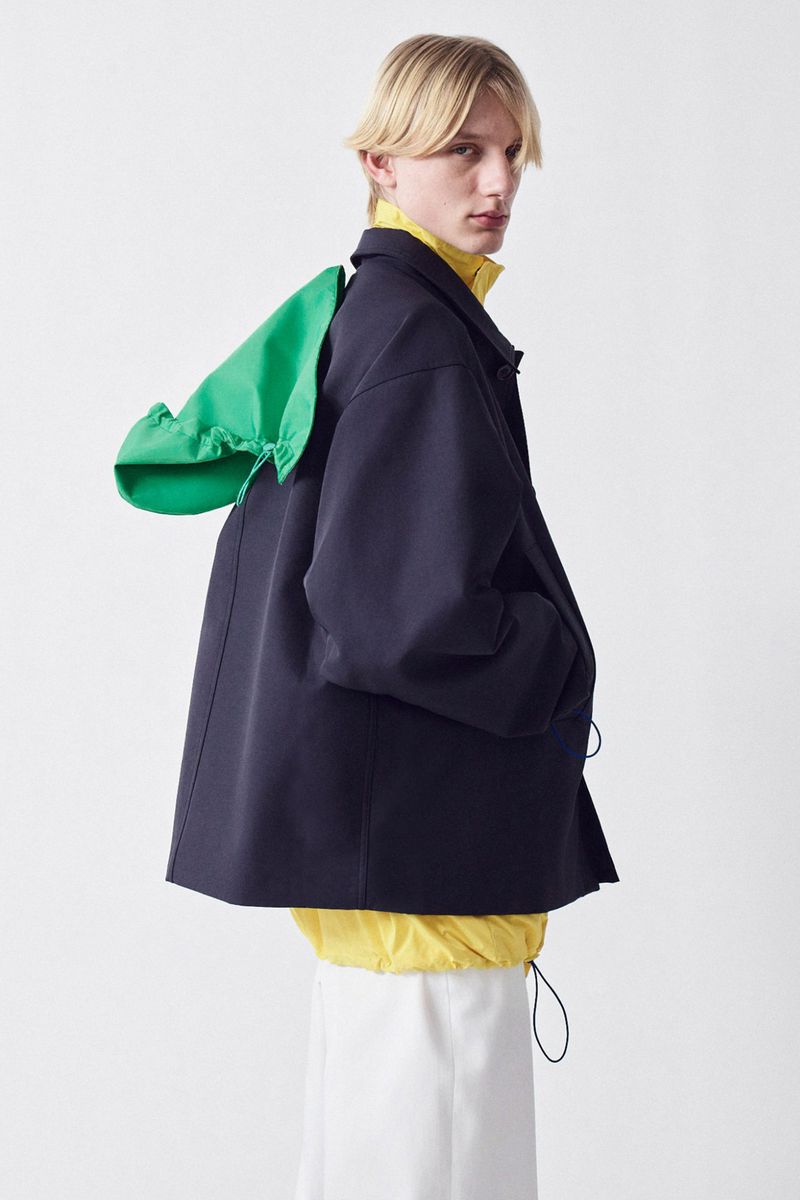 Male model in navy jacket with green hood, yellow turtleneck, and white skirt posing in photo studio against a white backdrop.