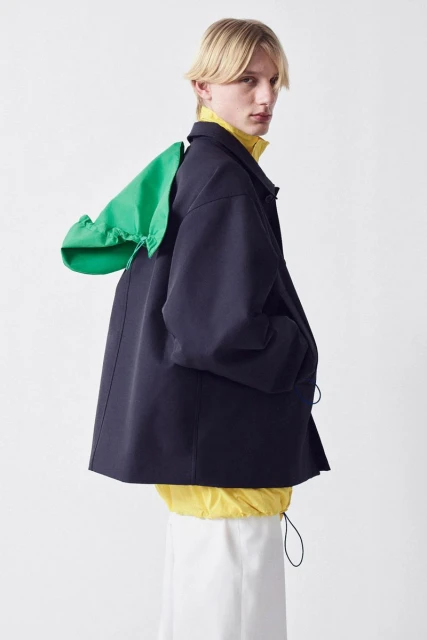 Male model in navy jacket with green hood, yellow turtleneck, and white skirt posing in photo studio against a white backdrop.