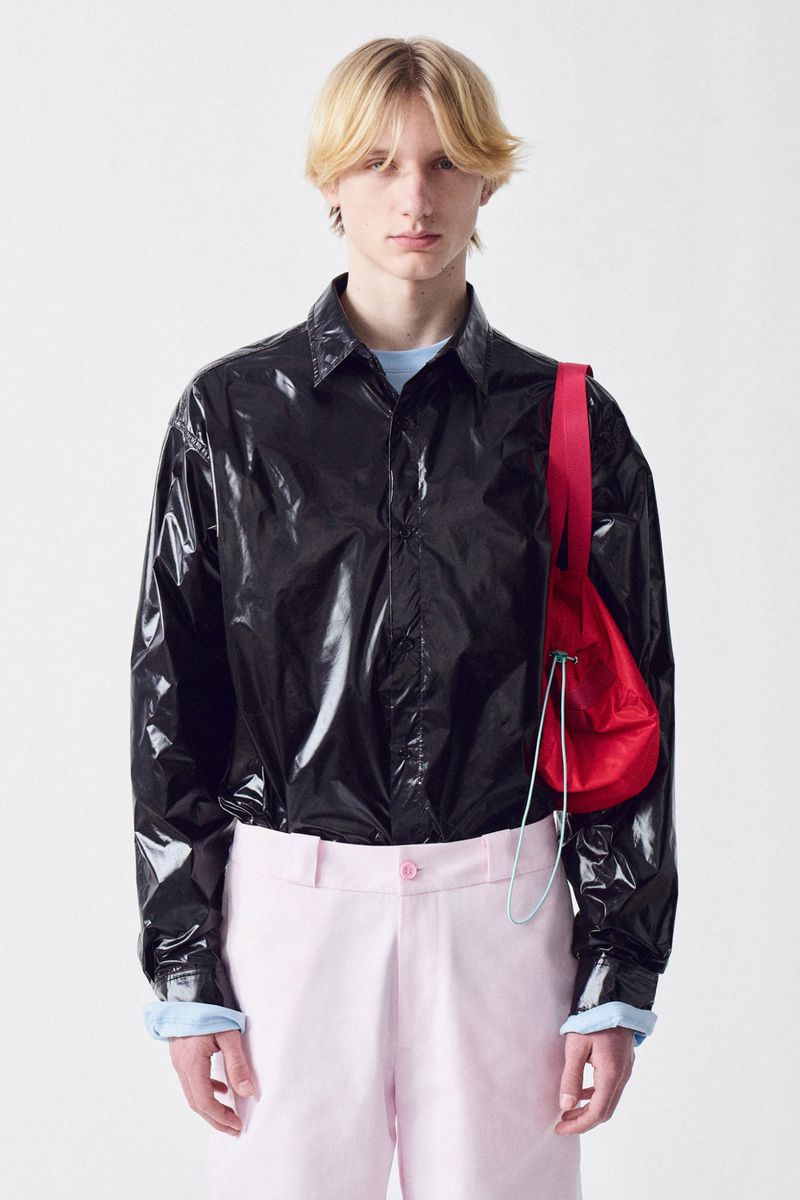 Young adult man poses indoors in a black shirt, pink pants, and a red bag on a white background in a fashion style portrait.