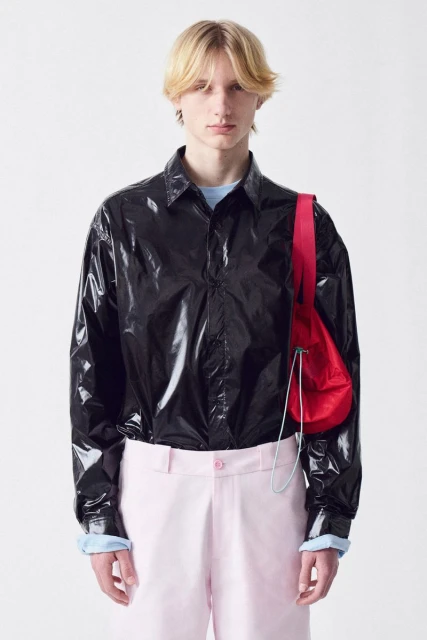 Young adult man poses indoors in a black shirt, pink pants, and a red bag on a white background in a fashion style portrait.
