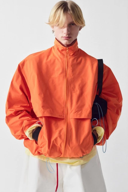 Young adult with blond hair in an orange zip-up jacket and white shorts standing in front of a white background in a studio shot.