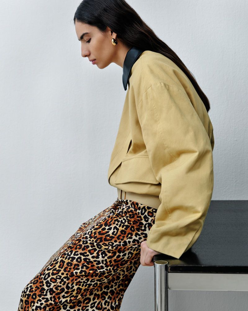 A young adult woman in a tan jacket and leopard print pants sits indoors next to a black table, looking down and to the left.