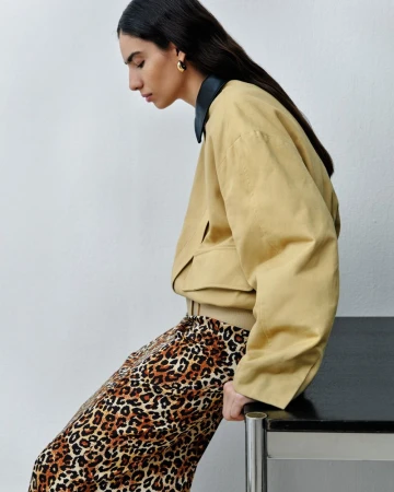 A young adult woman in a tan jacket and leopard print pants sits indoors next to a black table, looking down and to the left.