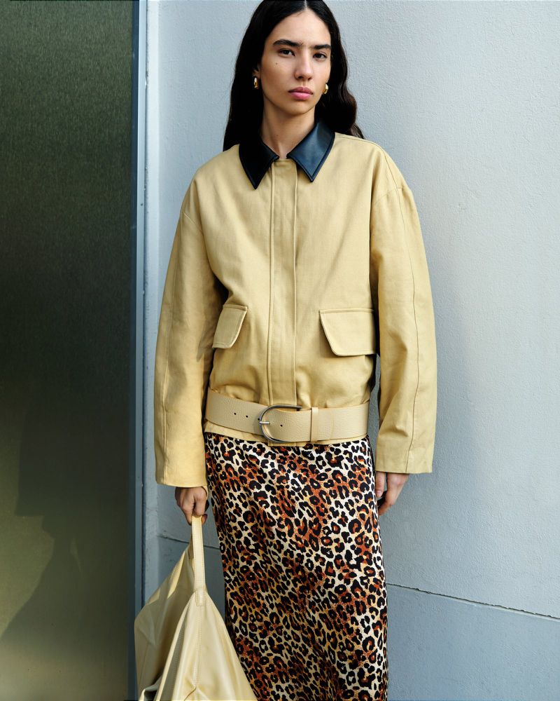 Woman in tan jacket and leopard print skirt holding tote bag stands against a grey wall with natural light. Fashion editorial.