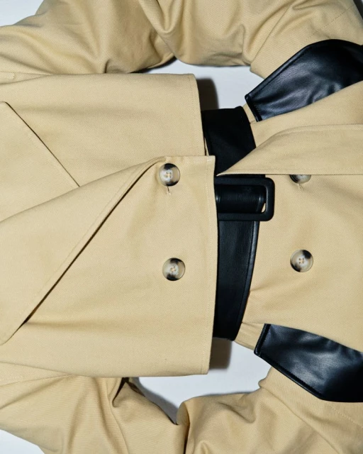 Tan trench coat with a black belt on a white background featuring neutral tones and classic fashion style.