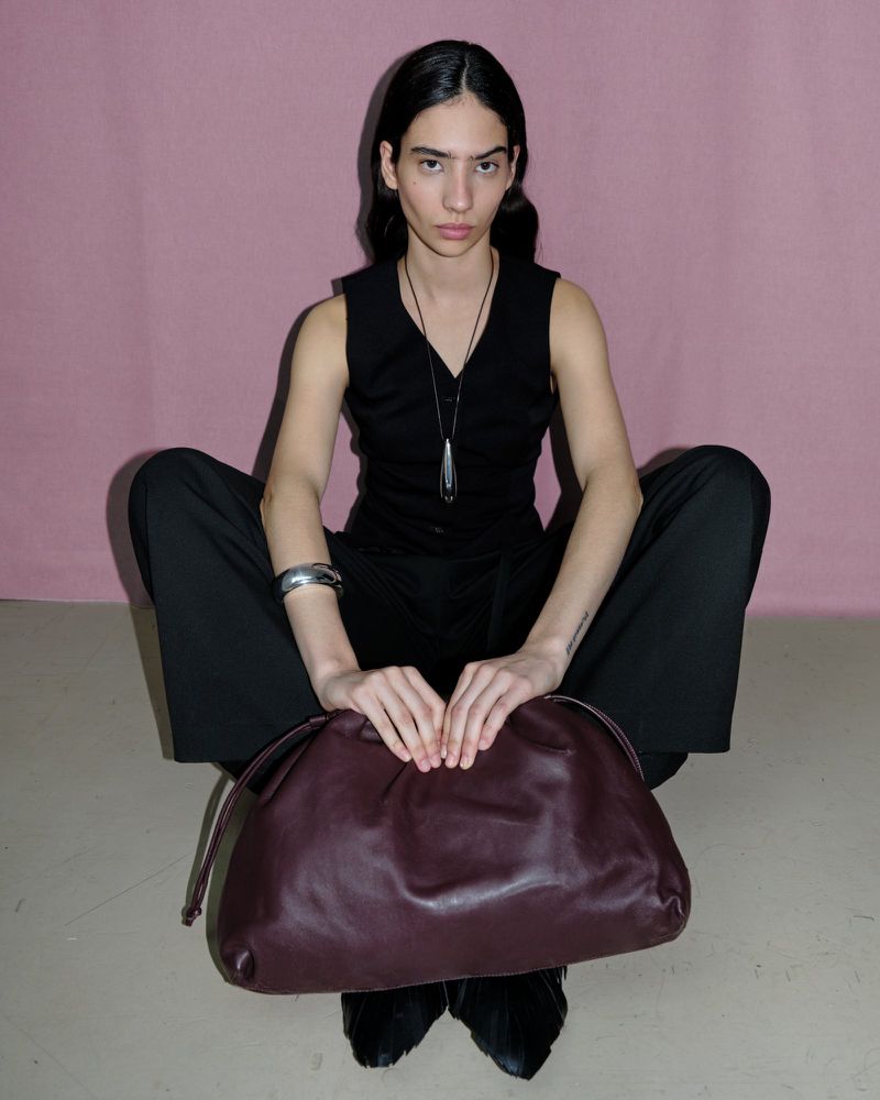 Model seated wearing a black vest and trousers while holding a dark leather clutch bag against a pink backdrop.