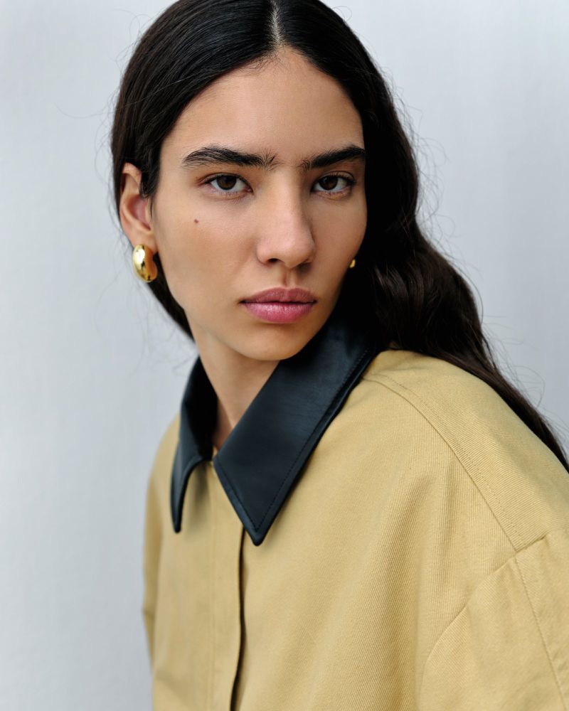 Portrait of a woman with dark hair wearing a tan shirt with a black collar and gold hoop earrings against a light grey background.