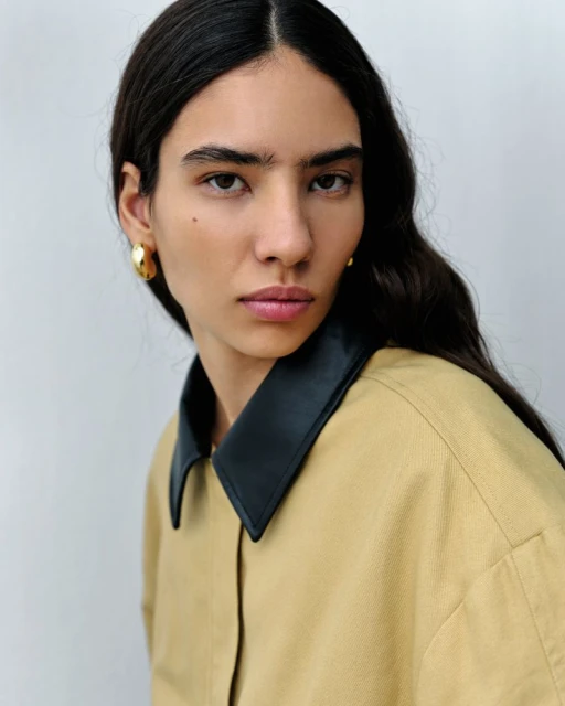 Portrait of a woman with dark hair wearing a tan shirt with a black collar and gold hoop earrings against a light grey background.