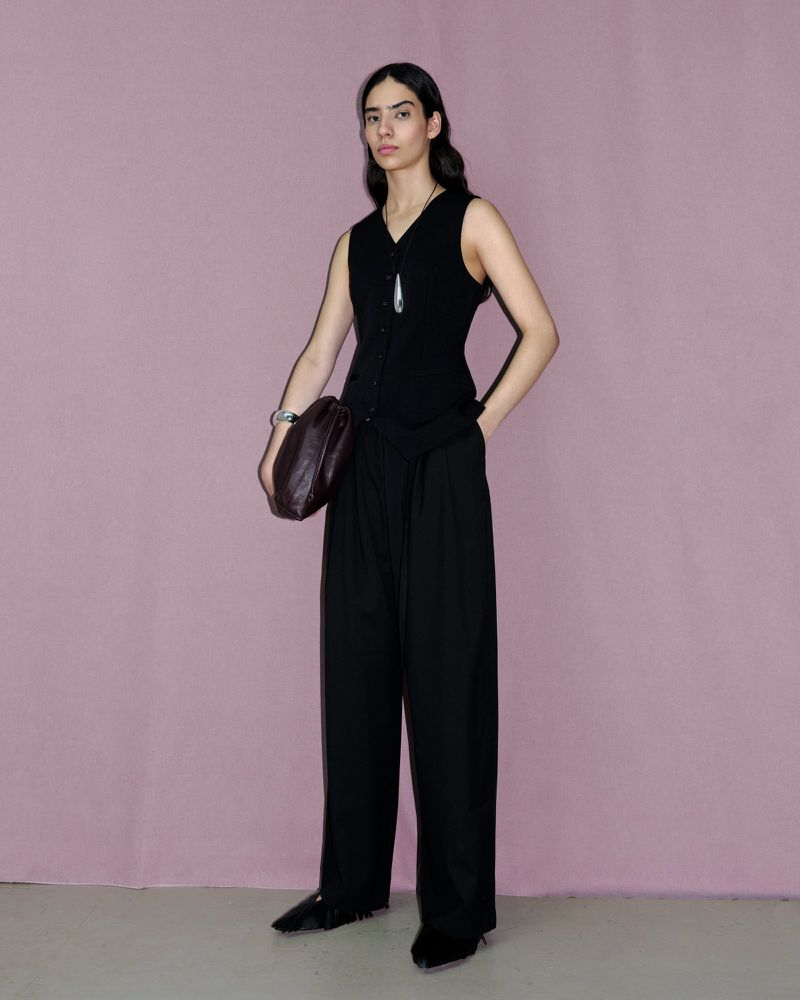 Full-length portrait of a model in a black vest and trousers holding a dark handbag against a pink background