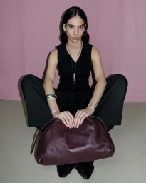 Model seated wearing a black vest and trousers while holding a dark leather clutch bag against a pink backdrop.