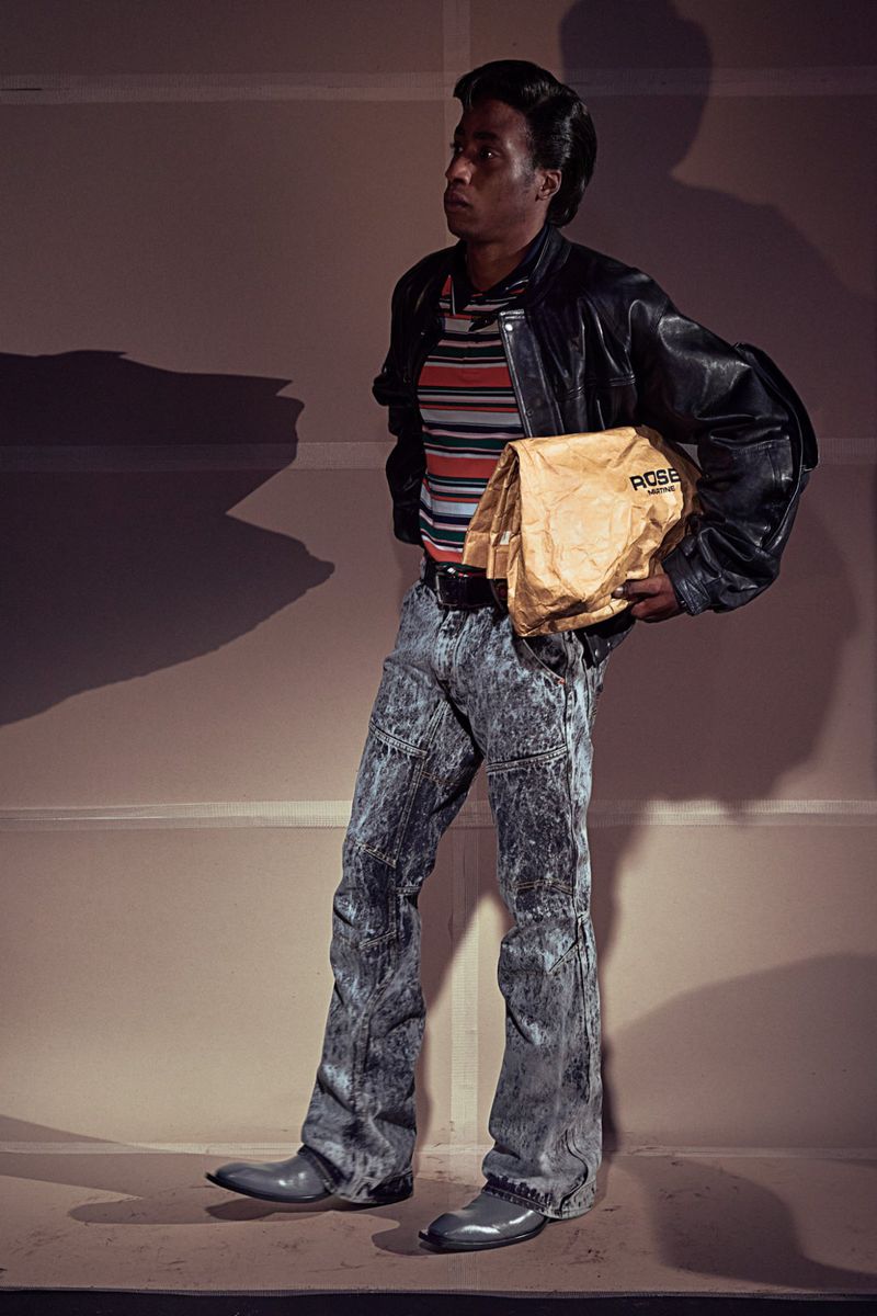 Adult male in retro clothing poses in a studio, holding a tan paper bag, featuring a black leather jacket, acid-wash jeans, and gray shoes.