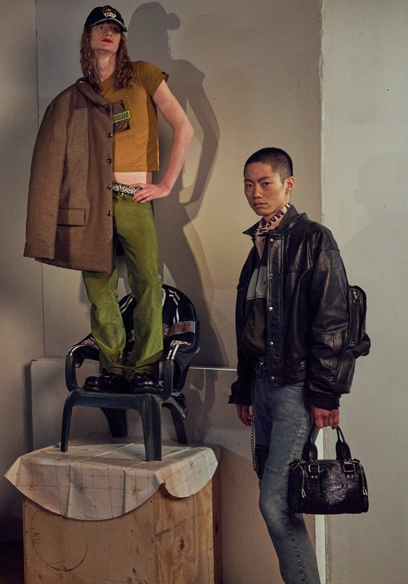 Two fashion models pose in front of a beige wall in a studio. One model is standing on a chair in green pants and a tan T-shirt.