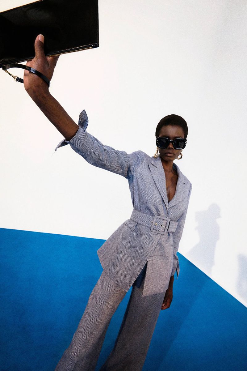 A fashion portrait features a woman in a gray suit and sunglasses holding a black purse against a blue and white graphic backdrop.