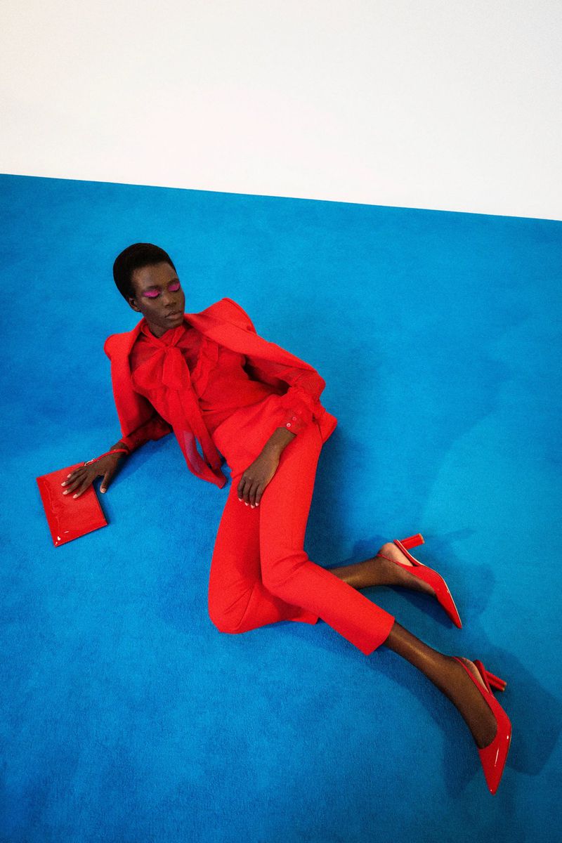 Fashionable woman in red suit and heels poses with clutch on blue. High-angle shot showcases studio fashion and vibrant color contrast.