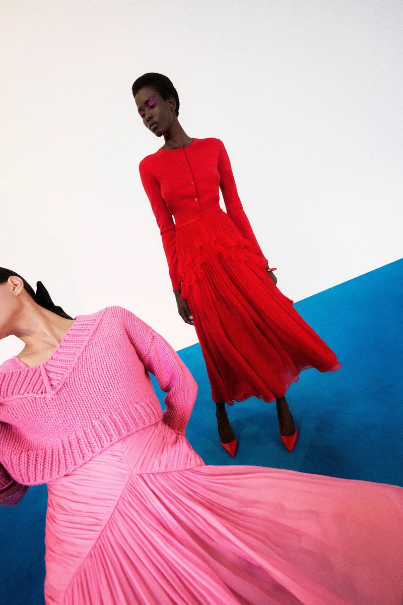 Two models pose indoors against a color-blocked backdrop wearing vibrant red and pink knitwear and pleated skirts for a fashion editorial.
