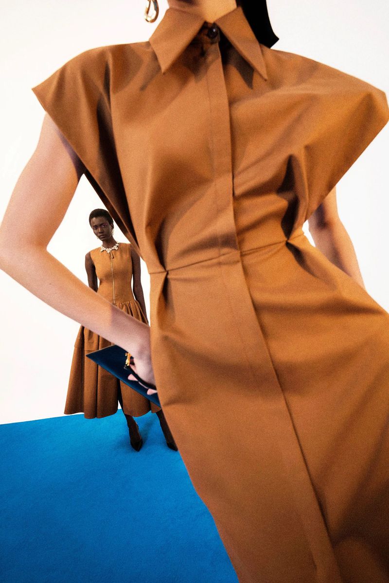 Two women in a fashion shot, both wearing brown sleeveless dresses, one holding a clutch, on a blue floor against a white background.