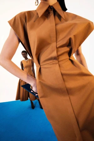 Two women in a fashion shot, both wearing brown sleeveless dresses, one holding a clutch, on a blue floor against a white background.