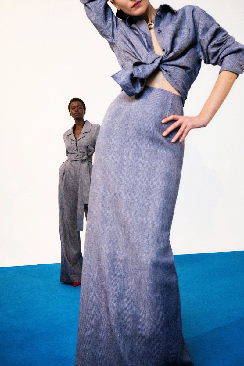 Two female models posing against a white wall with a bright blue floor, wearing matching denim-like outfits by Carolina Herrera.