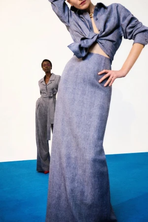 Two female models posing against a white wall with a bright blue floor, wearing matching denim-like outfits by Carolina Herrera.