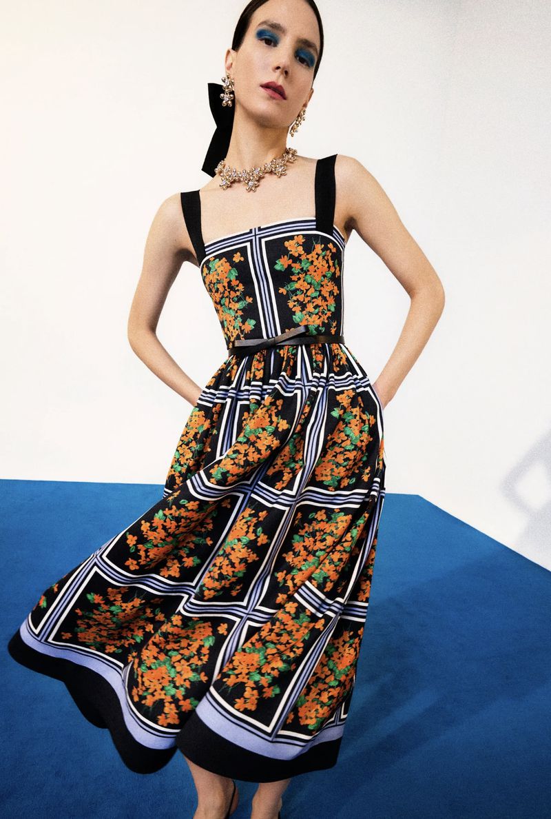 A woman poses in a black and orange floral print sundress with black straps and a black bow in her hair, against a white backdrop.