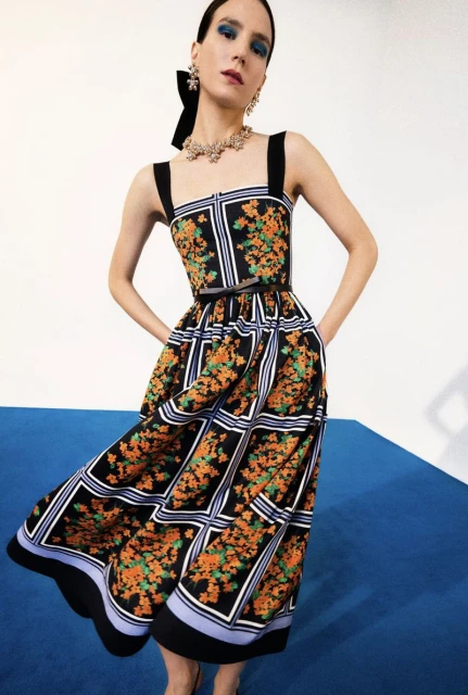 A woman poses in a black and orange floral print sundress with black straps and a black bow in her hair, against a white backdrop.