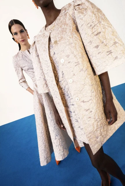 Two women in patterned formal wear in front of a blue and white background. The woman in front wears a coat and the other a dress suit.