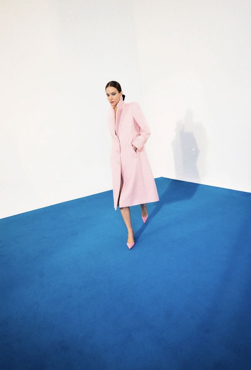 Fashion shot of a woman in a pink coat and matching heels standing on a blue platform against a white background in a photo studio.