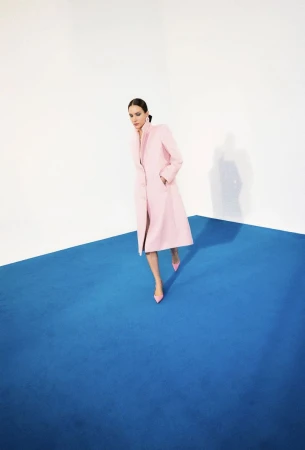Fashion shot of a woman in a pink coat and matching heels standing on a blue platform against a white background in a photo studio.