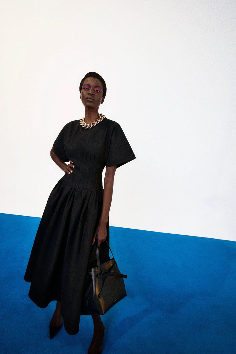 Model in black dress with chunky gold necklace holding a black handbag against a white and blue studio background