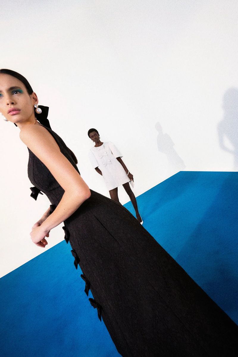 Model in a long black dress with bow details standing on a blue floor against a white wall for Carolina Herrera Pre-Fall 2025.