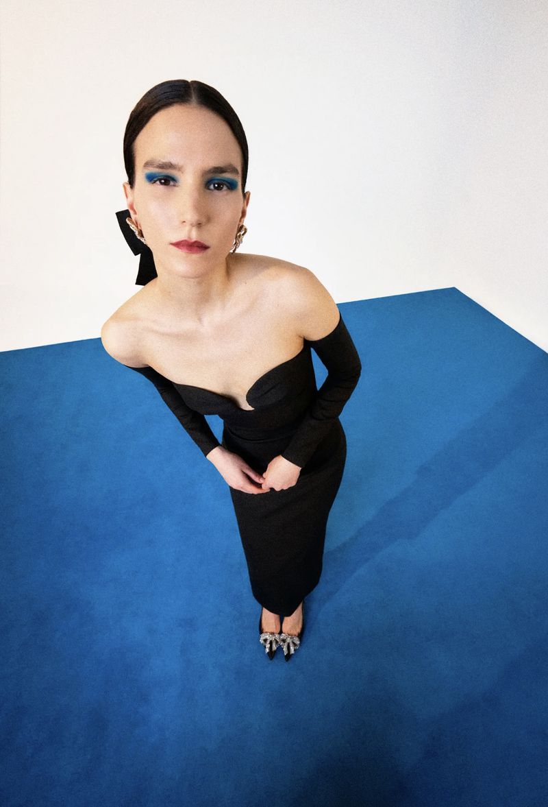 Woman in a long-sleeved black dress and embellished heels strikes a pose in a high-angle shot against a blue and white background.