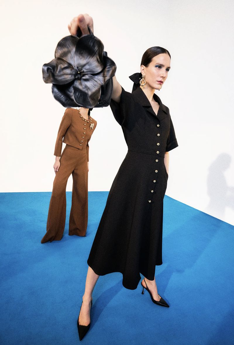 Two women pose in a studio with a black dress and a brown suit. One woman holds a large, black flower-shaped purse.