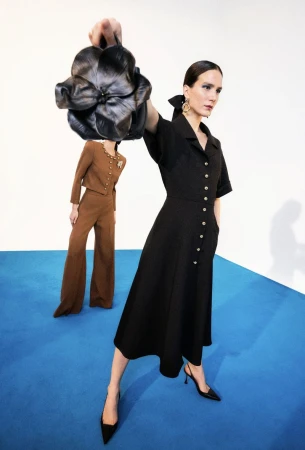 Two women pose in a studio with a black dress and a brown suit. One woman holds a large, black flower-shaped purse.