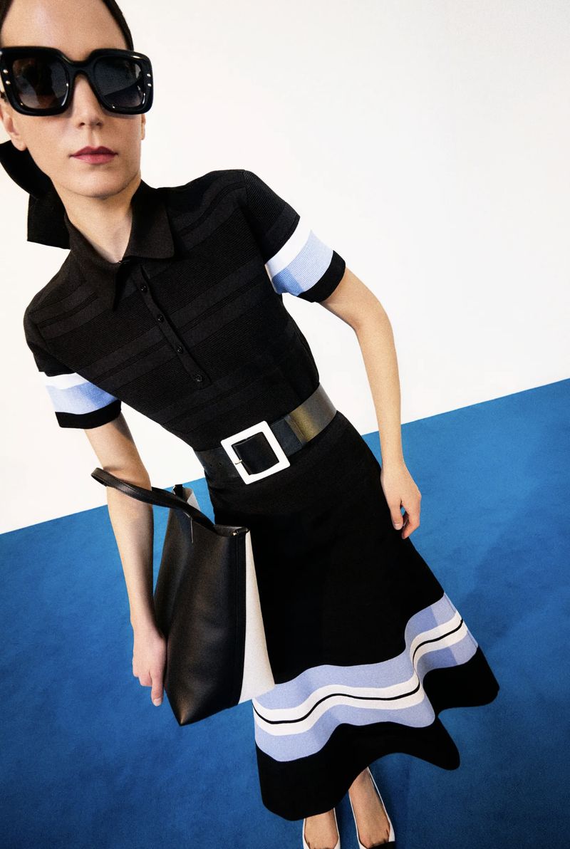 A woman poses in a black polo dress with a white-buckled belt, black tote bag, and sunglasses against a blue-and-white studio backdrop.