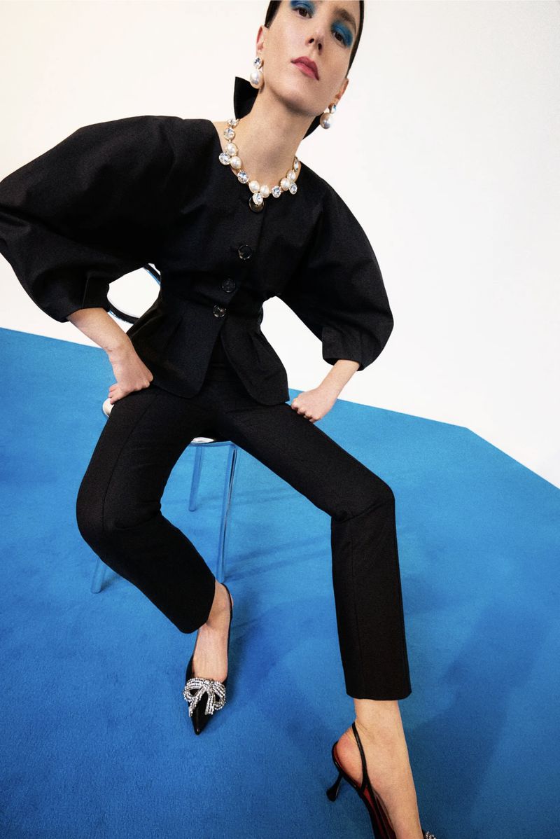 An adult female fashion model sits on a stool in an indoor photo studio, wearing a black two-piece suit, pumps, and pearl jewelry.