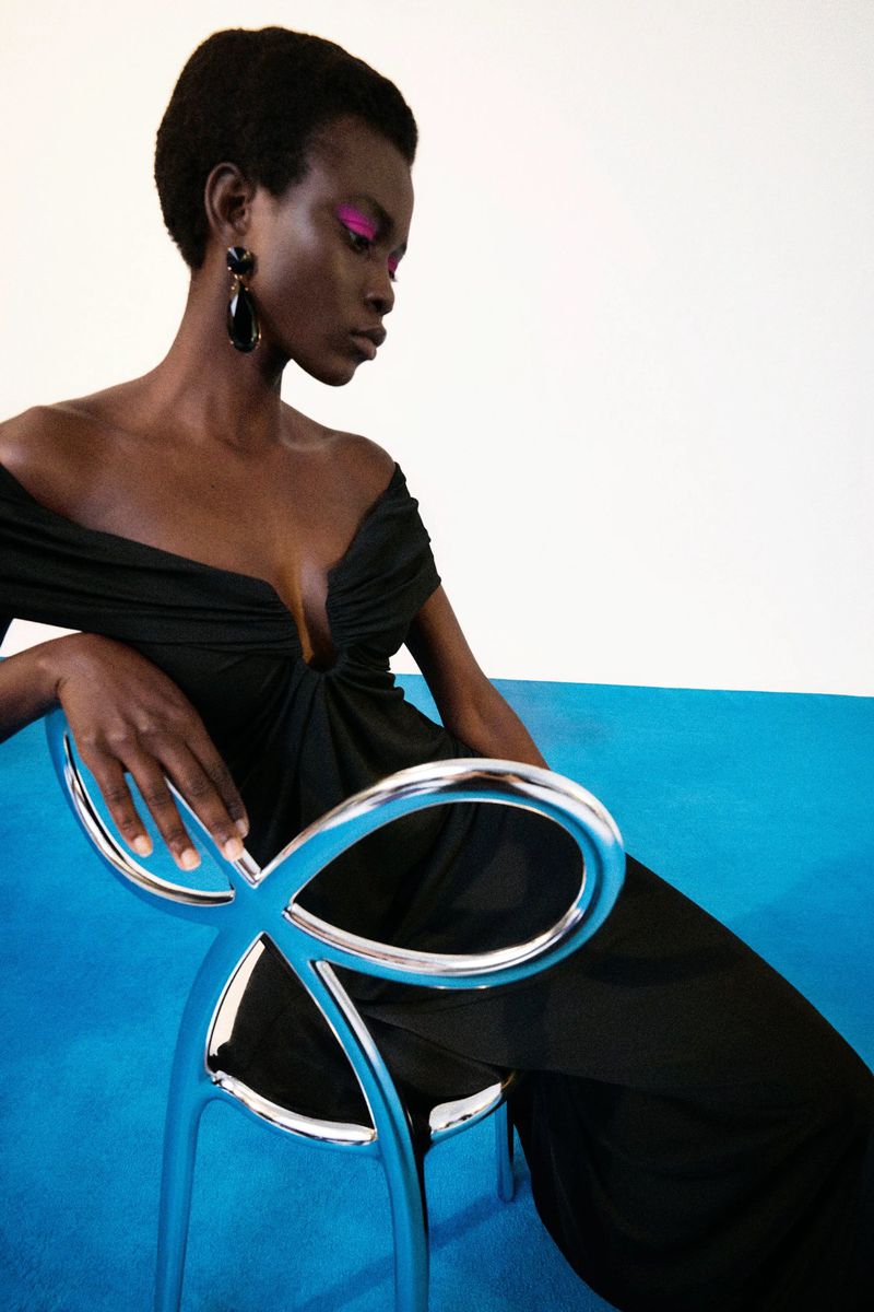 Woman with magenta eye makeup sits in a chrome chair wearing a black off-the-shoulder dress in a studio with a blue floor.