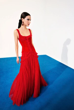 Model in a vibrant red ruffled gown standing on a blue carpet against a white studio wall