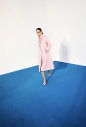 Model in pink coat walking across a vibrant blue floor against a plain white studio wall