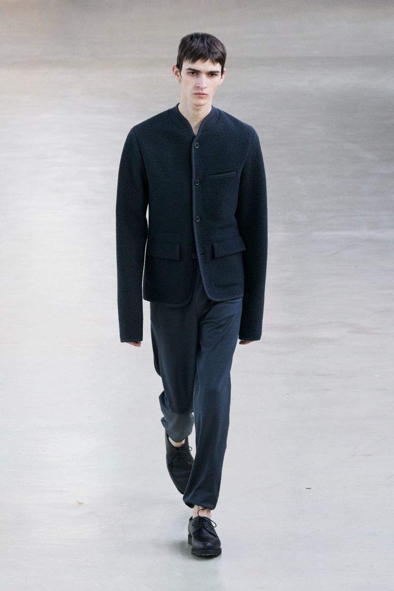 A young adult male model in a navy jacket and trousers walks on a bright, neutral-toned runway during a fashion show. Full shot.
