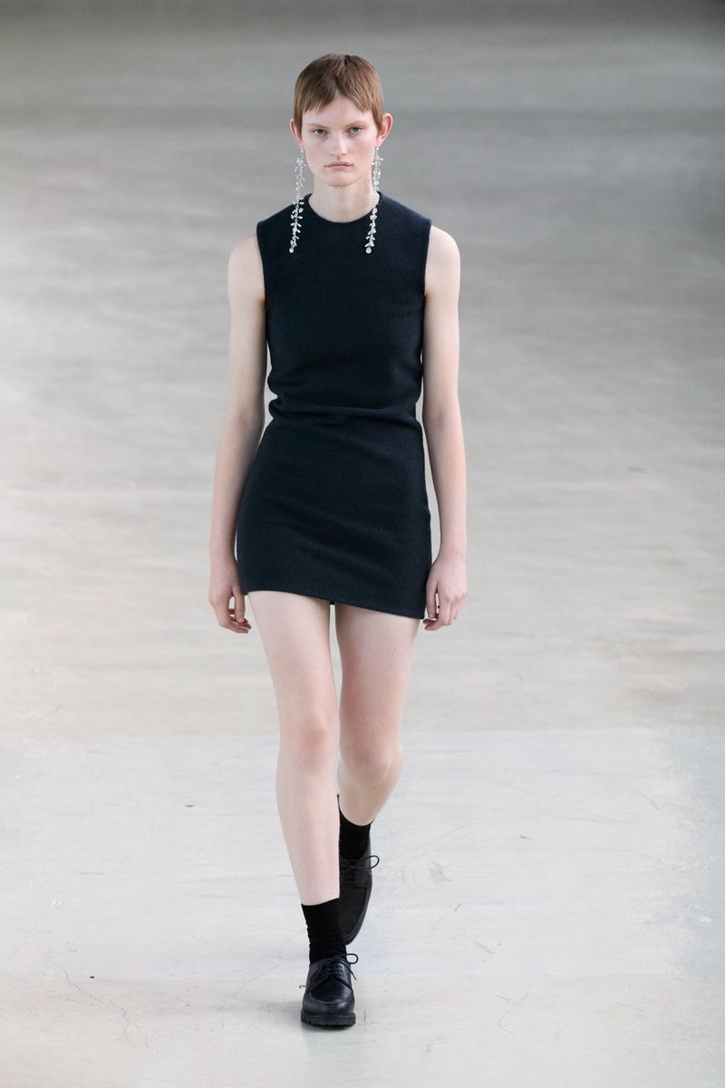 Model wearing a black sleeveless dress and drop earrings walking forward on a neutral floor