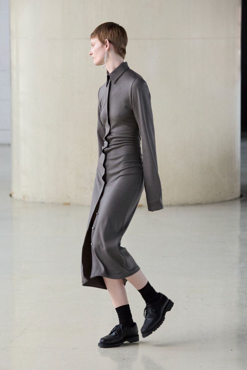A fair-skinned adult woman in a grey buttoned shirt-dress, black socks, and black leather shoes poses in a minimalist white photo studio.