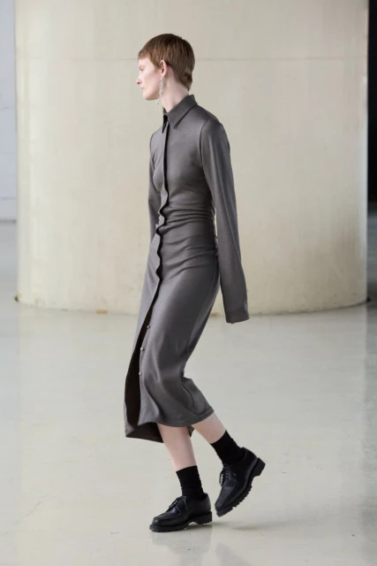 A fair-skinned adult woman in a grey buttoned shirt-dress, black socks, and black leather shoes poses in a minimalist white photo studio.