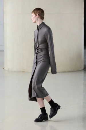 A fair-skinned adult woman in a grey buttoned shirt-dress, black socks, and black leather shoes poses in a minimalist white photo studio.