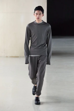 Asian male fashion model walks in a gray long-sleeve shirt, matching gray sweatpants, black beanie, and black leather shoes on a light gray floor.