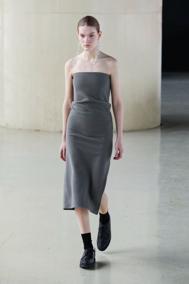 A slender woman walks the runway in a grey knit tube dress, black socks, and loafers. She wears long crystal earrings against a muted background.