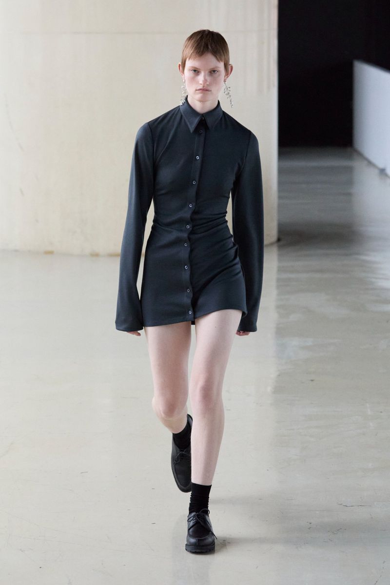 A model poses in a dark gray button-down mini dress, paired with black socks and flats, against a neutral concrete interior backdrop.