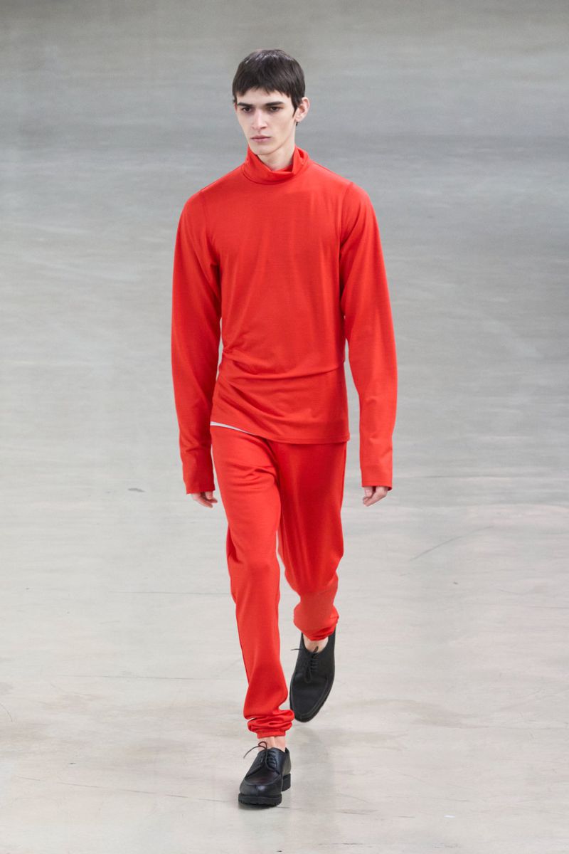 A male fashion model wearing a bright red turtleneck and matching pants walks a runway in a brightly lit fashion show venue.