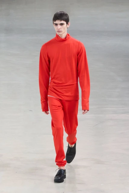 A male fashion model wearing a bright red turtleneck and matching pants walks a runway in a brightly lit fashion show venue.